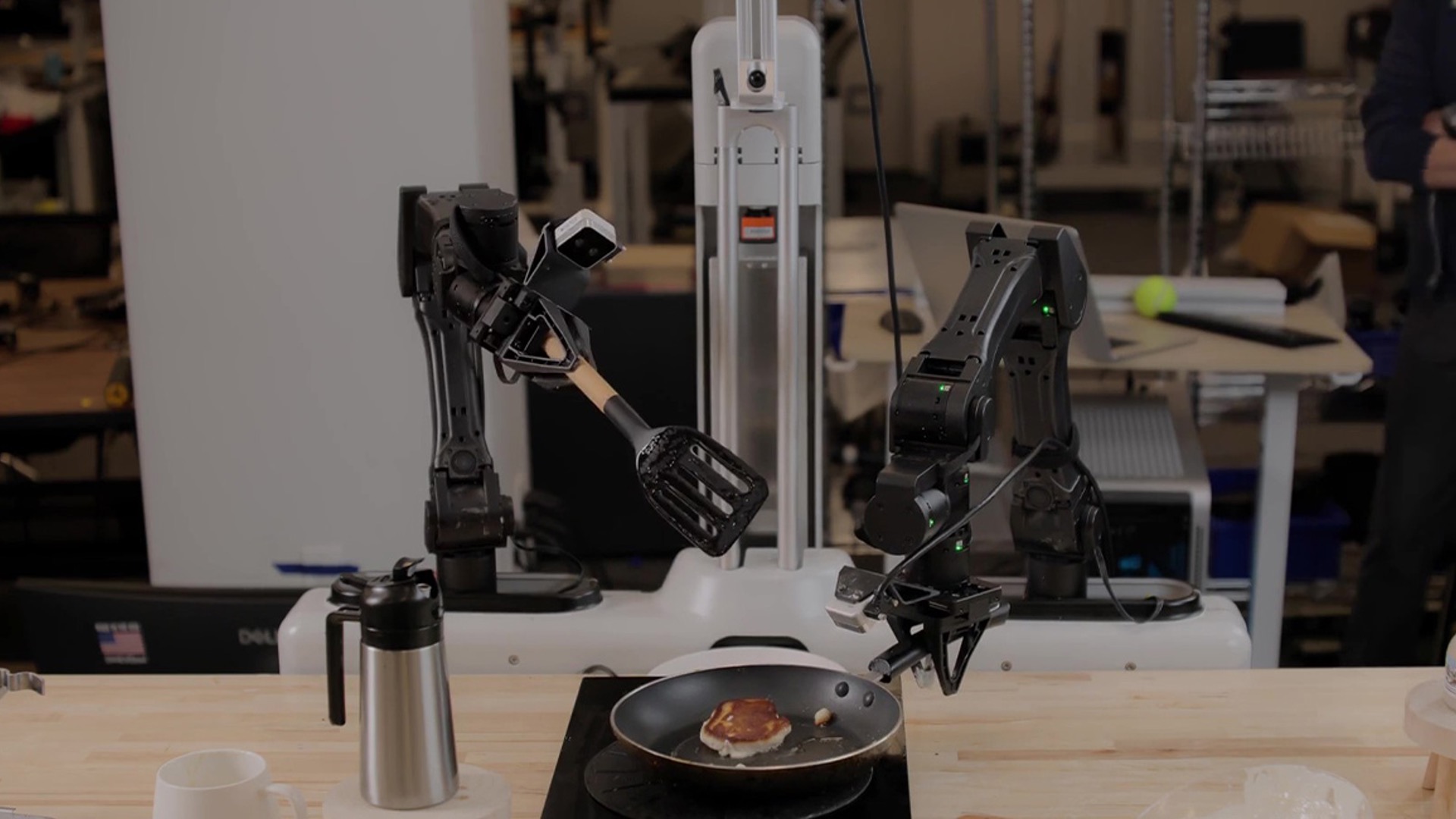 Le robot Skild équipé de deux bras articulés retourne une crêpe dans une poêle, sur un plan de travail de laboratoire